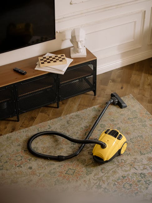 A yellow portable vacuum cleaner with a black hose and attached nozzle is positioned on a patterned area rug in a living room. The room features wooden flooring, white paneled walls, and a dark metal media console with a flat-screen TV mounted above. On top of the console, there are a remote control, a white book or magazine, a chessboard, and a small decorative sculpture. The scene is well-lit, showcasing a clean, tidy environment suitable for surface cleaning and maintenance, highlighting residential cleaning tasks that Tufnell Park Carpet Cleaning offers.