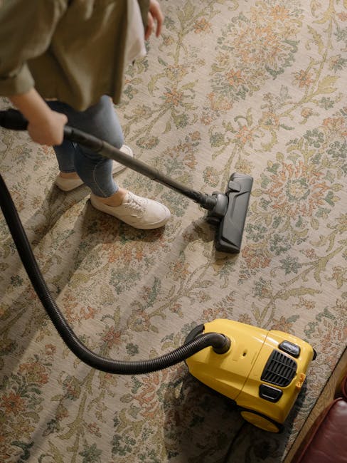 A person, dressed casually in a beige sweater, blue jeans, and white sneakers, is using a vacuum cleaner with a yellow body and black hose attachment to deep clean a patterned area rug in a residential room. The rug features an intricate floral design in muted tones of beige, green, and orange. The vacuum's hose is connected to a cleaning head that is actively engaged with the rug's surface, which appears clean and well-maintained. The room's lighting is warm and natural, highlighting the detailed texture of the rug and the cleanliness of the space. Tufnell Park Carpet Cleaning provides expert surface cleaning and hygienic deep cleaning services to maintain our homes' freshness and hygiene.