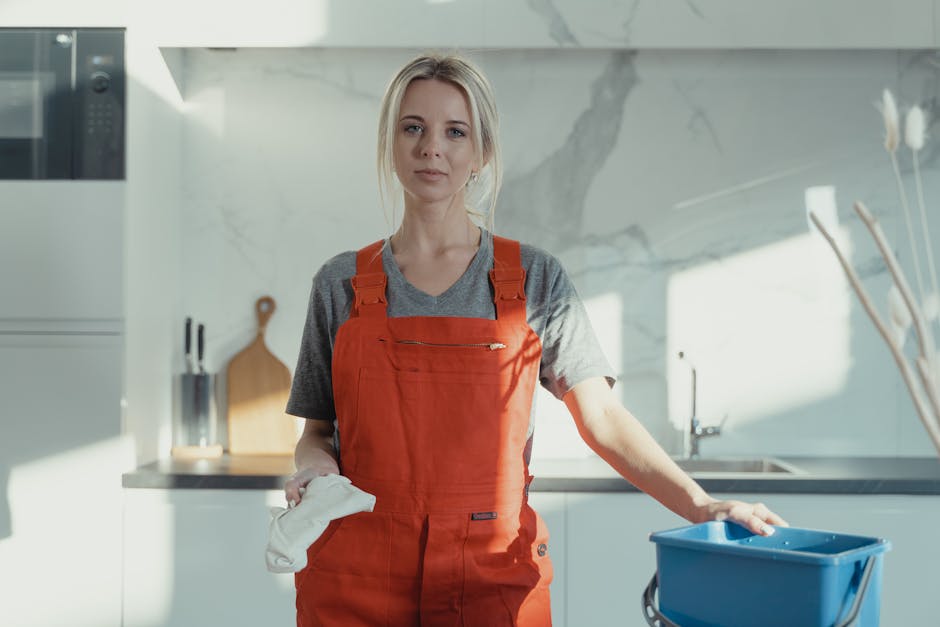A professional female cleaner with blonde hair tied back, dressed in a grey t-shirt and red cleaning overalls, standing in a modern kitchen. She holds a white cleaning cloth in her right hand and rests her left hand on a blue cleaning bucket. The kitchen features a white marble backsplash with grey veining, stainless steel appliances including an oven, and a sleek, minimalist countertop with a cutting board and knife block visible in the background. Bright natural light illuminates the space, highlighting the pristine and well-maintained surfaces, reflecting a focus on surface cleaning, hygiene, and domestic cleaning practices. The overall scene emphasizes thorough deep cleaning and sanitisation, aligning with the services provided by Tufnell Park Carpet Cleaning, as referenced on the page about avoiding hidden charges during cleaning bookings.