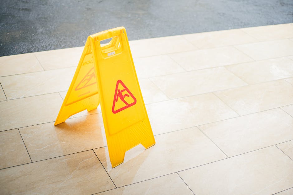 A bright yellow wet floor caution sign with a red triangular warning symbol stands on a tiled indoor floor, indicating a recently cleaned or wet surface. The floor consists of large, cream-colored ceramic tiles with visible grout lines, appearing clean and polished with a slight shine. The setting is well-lit, showing smooth, reflective surfaces and a neutral-toned space. In the background, part of the tiled floor continues, suggesting a corridor or entrance area. The image emphasizes the importance of surface cleaning, hygiene, and safety measures typical of professional domestic and commercial cleaning services provided by Tufnell Park Carpet Cleaning, ensuring environments are sanitized and hazards are minimized.
