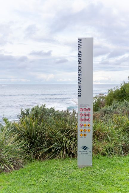 A tall, vertical signpost situated on a grassy area near the seaside, with ocean waves visible in the background under a partly cloudy sky. The signpost displays the name 'Malabar Ocean Pool' in large black letters at the top, followed by various safety and hygiene symbols, including no swimming, no diving, and other prohibitive icons. Below the symbols, the sign indicates the involvement of 'Rendellik City Council'. The surrounding scenery includes green shrubs and coastal vegetation, emphasizing a clean, well-maintained outdoor environment suitable for recreational use. This setting relates to cleanliness and safety guidelines for outdoor ocean pools, as outlined by local council regulations.
