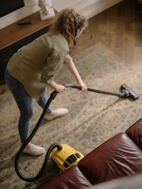 A woman with curly hair wearing a beige jacket, jeans, and white sneakers is engaged in deep cleaning a patterned residential carpet using a yellow vacuum cleaner with a long hose and nozzle. The room features wooden flooring with a dark finish, and a large area rug with floral motifs in beige, blue, and pink tones covers part of the floor. She is positioned near a dark red leather sofa, and a wooden sideboard with a white decorative item is visible against the wall. Bright natural light illuminates the space, highlighting the cleanliness of the area, and demonstrating surface cleaning and maintenance typical of domestic hygiene practices. Tufnell Park Carpet Cleaning offers professional deep cleaning services for carpets in Tufnell Park N19, ensuring hygienic and well-maintained environments.