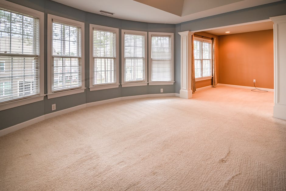 A spacious living room with a U-shaped arrangement of large double-hung windows featuring white wooden blinds, allowing natural light to brighten the space. The walls are painted in soft muted tones—light gray in the main area and a warm beige accent wall—enhanced by white trim and decorative columns framing the transition between rooms. The beige carpeted floor appears clean and plush, with subtle indentations from foot traffic. The room includes a recessed ceiling with concealed lighting, and a white baseboard runs along the perimeter. Visible electrical outlets are located along the walls. The image showcases an immaculate, well-maintained interior ideal for professional surface cleaning and house sanitisation by Tufnell Park Carpet Cleaning, emphasizing the importance of deep cleaning for hygiene and maintenance of domestic spaces.