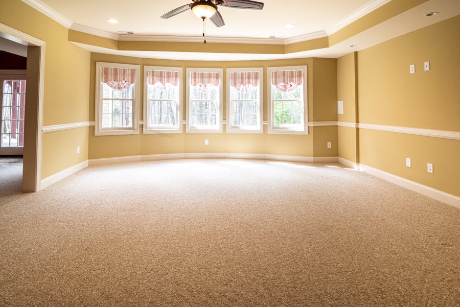 A woman with curly hair wearing a beige jacket, jeans, and white sneakers is engaged in deep cleaning a patterned residential carpet using a yellow vacuum cleaner with a long hose and nozzle. The room features wooden flooring with a dark finish, and a large area rug with floral motifs in beige, blue, and pink tones covers part of the floor. She is positioned near a dark red leather sofa, and a wooden sideboard with a white decorative item is visible against the wall. Bright natural light illuminates the space, highlighting the cleanliness of the area, and demonstrating surface cleaning and maintenance typical of domestic hygiene practices. Tufnell Park Carpet Cleaning offers professional deep cleaning services for carpets in Tufnell Park N19, ensuring hygienic and well-maintained environments.